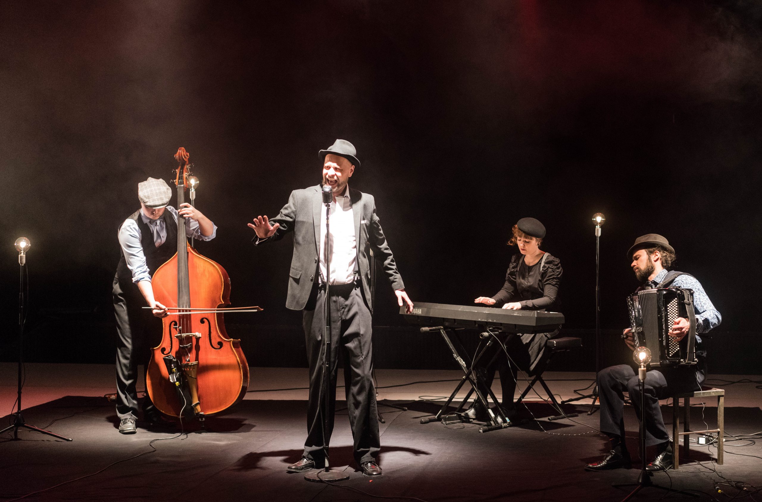 kolme muusikkoa ja yksi laulaja. vasemmalla basisti, keskellä takana pianisti, oikealla haitaristi ja keskellä edessä laulaja laulaa. kaikki ovat pukeutuneet 30-luvun vaatteisiin.